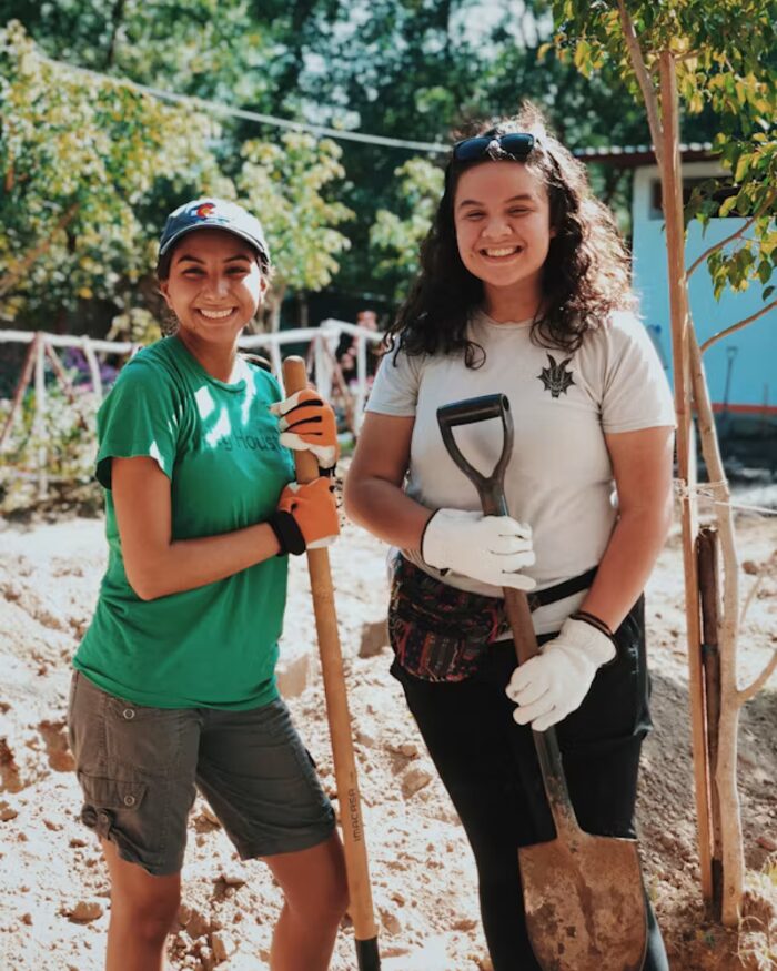 Two smiling Community First Alliance volunteers planting a young tree together in warm afternoon sun